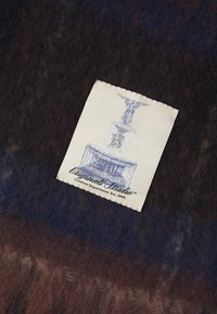 Wollen stof met een donkerbruine en blauwe gradient. Een crèmekleurig label heeft een blauwe illustratie van een monument en de tekst "Originals Studio."