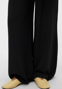 Czarne, szerokie spodnie o fakturowanej tkaninie, zestawione z żółtymi, płaskimi butami o zaokrąglonym nosku i subtelnych wzorach.