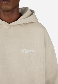 Bēšas kapuces džemperis ar balto "Pegador" izšūto logotipu uz krūtīm. Mīksta auduma, ar brīvu piegriezumu un ribota struktūra.