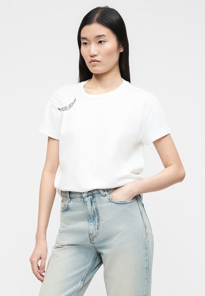 Νεαρή γυναίκα με μακριά μαύρα μαλλιά φοράει λευκό μπλουζάκι με μικρό σχέδιο φτερού και ανοιχτό μπλε ψηλόμεσο τζιν, με το χέρι στην τσέπη.