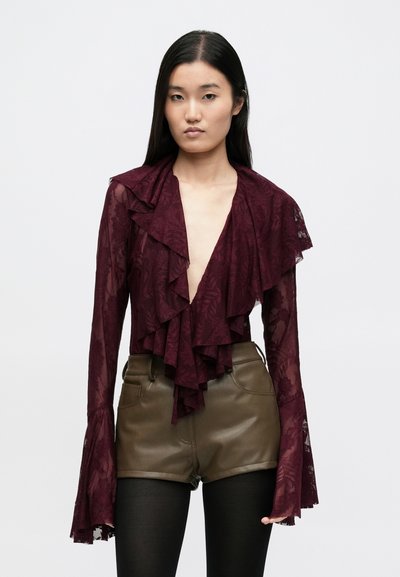 Haut en dentelle bordeaux avec froufrous à l'avant, décolleté en V profond et manches évasées. Associé à un short en cuir vert olive et des collants noirs.