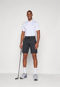 Άσπρο πόλο μπλουζάκι με γιακά, μαύρο σορτς, γκρι αθλητικά παπούτσια και μια ράβδο γκολφ, στέκεται μπροστά σε ένα απλό φόντο.