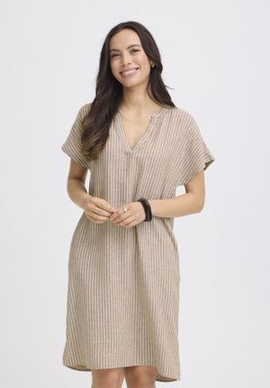 Usmievajúca sa žena s dlhými tmavými vlasmi, oblečená v béžovo-bielych pásikavých krátkych šatách a čiernom náramku, stojaca pred jednoduchým pozadím.