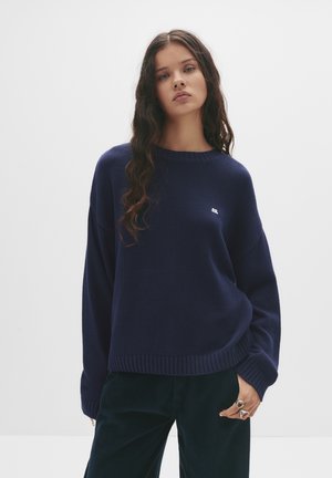 Pulover tricotat de culoare bleumarin, cu margini și manșete cu model côtelé, având un logo mic pe față. Textură moale și croială lejeră.