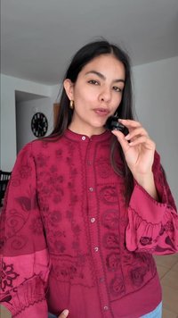 Γυναίκα με κόκκινο πουκάμισο με λουλούδια κρατάει μια μαύρη βούρτσα μακιγιάζ κοντά στα χείλη της σε έναν μοντέρνο χώρο με ένα ρολόι στον τοίχο.