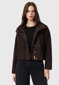 Giacca corta in camoscio marrone con collo alto, dettagli in oro, chiusura con zip e bottoni a pressione. Caratterizzata da una vestibilità rilassata e una texture morbida.