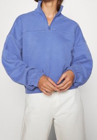 Suéter azul claro con cuello de medio cierre, puños elásticos y un corte relajado. Tejido suave con detalle de costura visible.