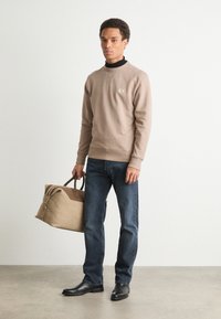 Beige sweatshirt med et lille logo, mørke jeans og sorte lædersko. Bærer en tan duffel taske med brune detaljer. Enkelt, afslappet outfit.