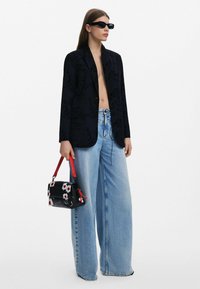 Svart kavaj med subtila grafiska mönster, högmidjade ljusblå vida jeans och en liten svart handväska med blommor och röd rem.