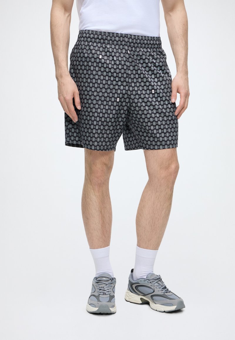 Homme portant un short noir à motifs, des chaussettes blanches, des baskets grises et un t-shirt blanc sur un fond uni.