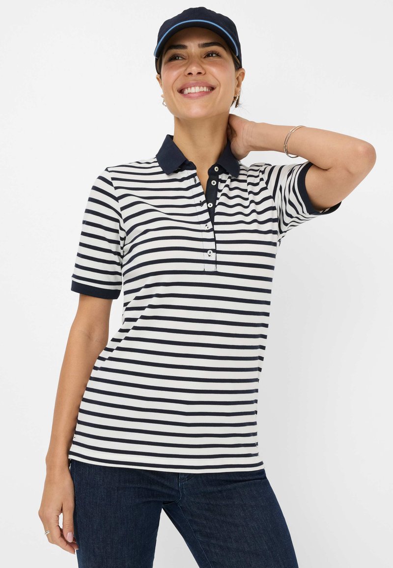 Polo a righe blu navy e bianche con maniche corte, caratterizzato da un colletto blu navy a tinta unita e un patta con quattro bottoni. I jeans in denim completano il look.