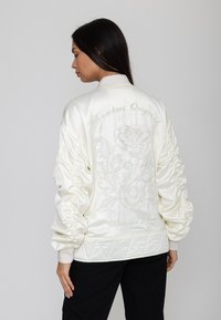 Femme aux longs cheveux foncés porte une veste bomber en satin blanc brodée d'un grand motif floral et d'un texte en cursive dans le dos.