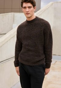 Bruine, textuurgebreide trui met een ronde hals en geribbelde manchetten, gecombineerd met zwarte broek. De stof lijkt zacht en fuzzy.