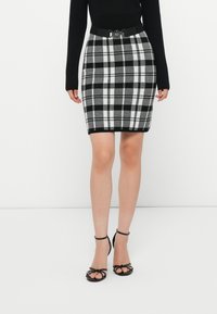 Μαύρη και άσπρη καρό μολυβένια φούστα με εφαρμοστό σχέδιο, μαύρη ζώνη με κυκλικό κούμπωμα, φορεμένη με μαύρα τακούνια με λουριά.