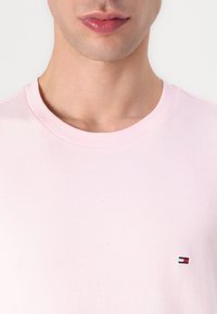 T-shirt en coton rose clair avec un col rond, comportant un petit logo brodé en rouge, blanc et bleu sur le côté gauche de la poitrine.