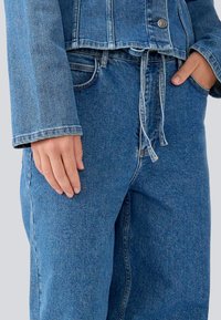 Chaqueta de mezclilla azul claro con un diseño cropped, con cierre de botones y mangas anchas. Combinada con pantalones anchos a juego y cordón.