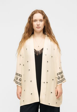 Jeune femme aux longs cheveux porte un kimono crème avec des imprimés de palmiers noirs et des poignets à motifs sur un top en dentelle noire, debout sur un fond blanc.