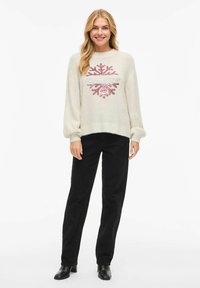 Kremo spalvos megztinis su minkšta tekstūra, puoštas rožinės spalvos snaigės dizainu ir užrašu „leisk snigti“, derinamas su juodomis tiesios kirpimo kelnėmis ir auliniais batais.