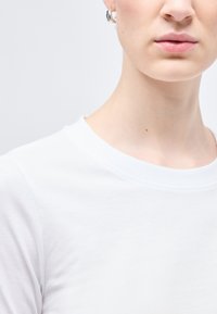 Κοντινό πλάνο του κάτω μέρους προσώπου και λαιμού ενός ατόμου που φοράει λευκό μπλουζάκι με στρογγυλή λαιμόκοψη  και ένα μικρό ασημένιο κρίκο στο αυτί, σε γκρίζο φόντο.