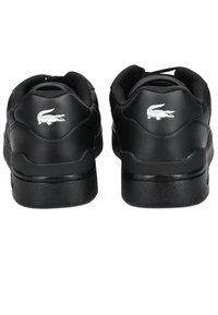 Black sneaker with smooth synthetic material, padded collar, and contrasting white crocodile logo on the heel. Flat rubber sole.