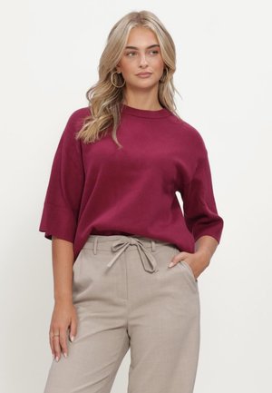 Donna con lunghi capelli biondi, che indossa grandi orecchini a cerchio, una maglia larga bordeaux con maniche fino al gomito e pantaloni beige con cintura da annodare, in piedi con una mano in tasca.