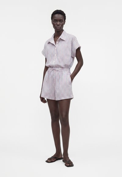 Γυναίκα που φορά κοντομάνικο ριγέ ολόσωμο με γιακά, ελαστική μέση και μαύρες σαγιονάρες, στέκεται σε λευκό φόντο.