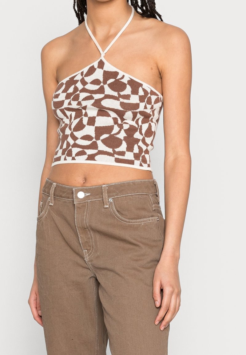 Femme portant un crop top dos nu à motif abstrait marron et blanc et un pantalon marron taille haute avec des coutures blanches sur fond uni.