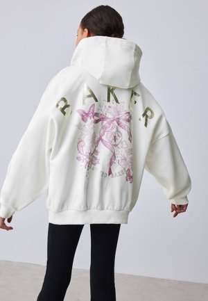 Osoba nosi preveliku bijelu hoodicu s kapuljačom, sa zlatnim tekstom "BAKER" i ružičastim cvjetnim i leptir dizajnom na leđima.