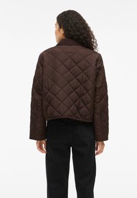 Mulher com cabelo encaracolado, vestindo uma jaqueta acolchoada castanha-escura e calças pretas, de pé, de costas, contra um fundo branco.