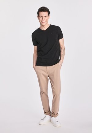 Czarny t-shirt z dekoltem w serek, beżowe spodnie i białe tenisówki. Ogólnie slim fit, miękka tkanina, krótkie rękawy, z widocznym tatuażem na ramieniu.