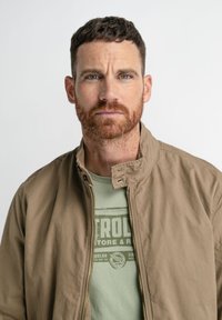 Chaqueta bomber marrón clara con cuello alzado, cierre de cremallera y bolsillos laterales, llevada sobre una camiseta gráfica de color verde claro. Textura de tela suave.