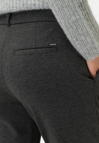 Harmaat kangashousut sisältävät takataskun, jossa on tyylikäs musta reunus ja pieni metallikoriste. Kankaan pinta on sileä ja hyvin muotoiltu.