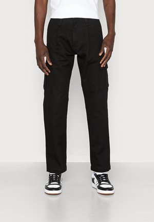 Front view of person wearing black cargo pants with side pockets and black-and-white sneakers, standing on a light floor.