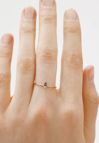 Anillo de oro con una banda delgada, que presenta una pequeña piedra verde y un diminuto acento de diamante, llevado en una mano parcialmente visible.
