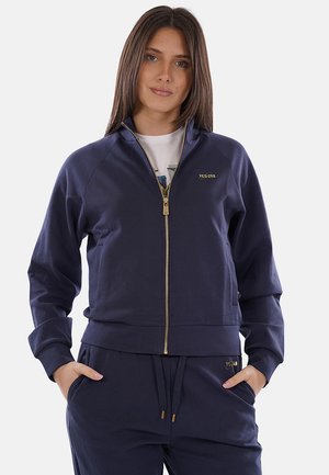 Donna che indossa una giacca blu scuro con zip e pantaloni abbinati, con le mani in tasca, in piedi davanti a uno sfondo chiaro e uniforme.