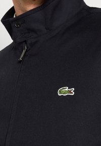 Granatowa kurtka z wysokim kołnierzem, z logo zielonego krokodyla obramowanym na biało po lewej stronie. Wykonana z gładkiego, lekkiego materiału.