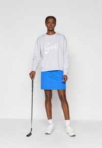 Grå hettegenser med teksten "Golf", blå skjørt, hvite sokker og hvite sko. Holder en golfklubbe, står på en ensfarget bakgrunn.