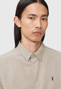 Camicia in corduroy di cotone beige con colletto a punta, chiusura frontale con bottoni e un accento ricamato a forma di teschio di capra sul lato sinistro.