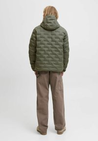 Osoba v olivově zelené prošívané bundě s kapucí a volných hnědých kalhotách, stojící čelem od sebe na jednobarevném bílém pozadí.