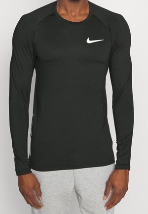 Osoba ubrana w czarną koszulkę Nike z długim rękawem z białym logo na klatce piersiowej oraz jasnoszare spodnie, stojąca na jednolitym tle.