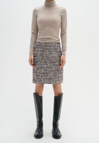 Suéter beige de cuello alto hecho de material suave, combinado con una falda lápiz de tweed texturizada y multicolor, y botas negras hasta la rodilla.