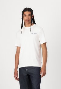 Hvid bomulds t-shirt med korte ærmer, med blå broderet logo på venstre bryst, parret med mørk denim jeans.