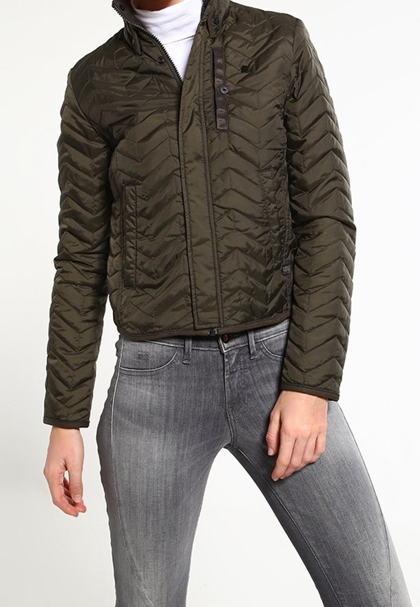 Veste matelassée vert olive avec motif en chevrons, fermeture éclair, col montant et subtils accents marron, portée avec un jean gris.