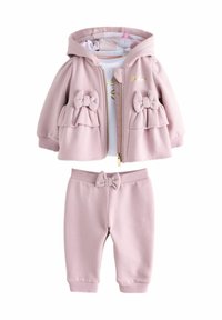 Béžová pink kapucňová bunda s detailmi mašle a zodpovedajúcimi elastickými nohavicami, doplnená o bielu košeľu s zlatým dizajnom.