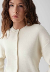 Crèmekleurige gebreide cardigan met een ronde halslijn, lange mouwen en vijf knopen. Het heeft een zachte textuur en lichte ribbeldetails.