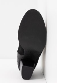 Semelle d'une botte noire à talon carré texturé, positionnée verticalement contre un mur d'angle blanc.