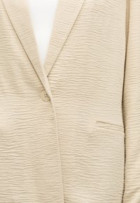 Μπεζ σακάκι με υφή, με ένα κουμπί κλεισίματος, πέτα γιακάδες με χαραγή και μπροστινή τσέπη με φάσα πάνω από λευκό τοπ.