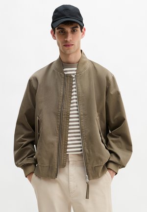 Young man wearing a black cap, olive bomber jacket over a beige and white striped shirt, with light beige pants, hands in pockets.