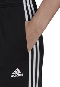 Černé sportovní kalhoty se třemi bílými pruhy. Mají boční kapsu a bílé logo Adidas. Vyrobeno z hladkého materiálu.