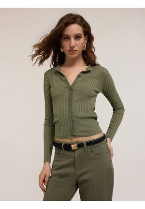 Green ribbed cardigan with a collared neckline and button front. Worn with olive green pants and a black belt featuring a gold buckle.